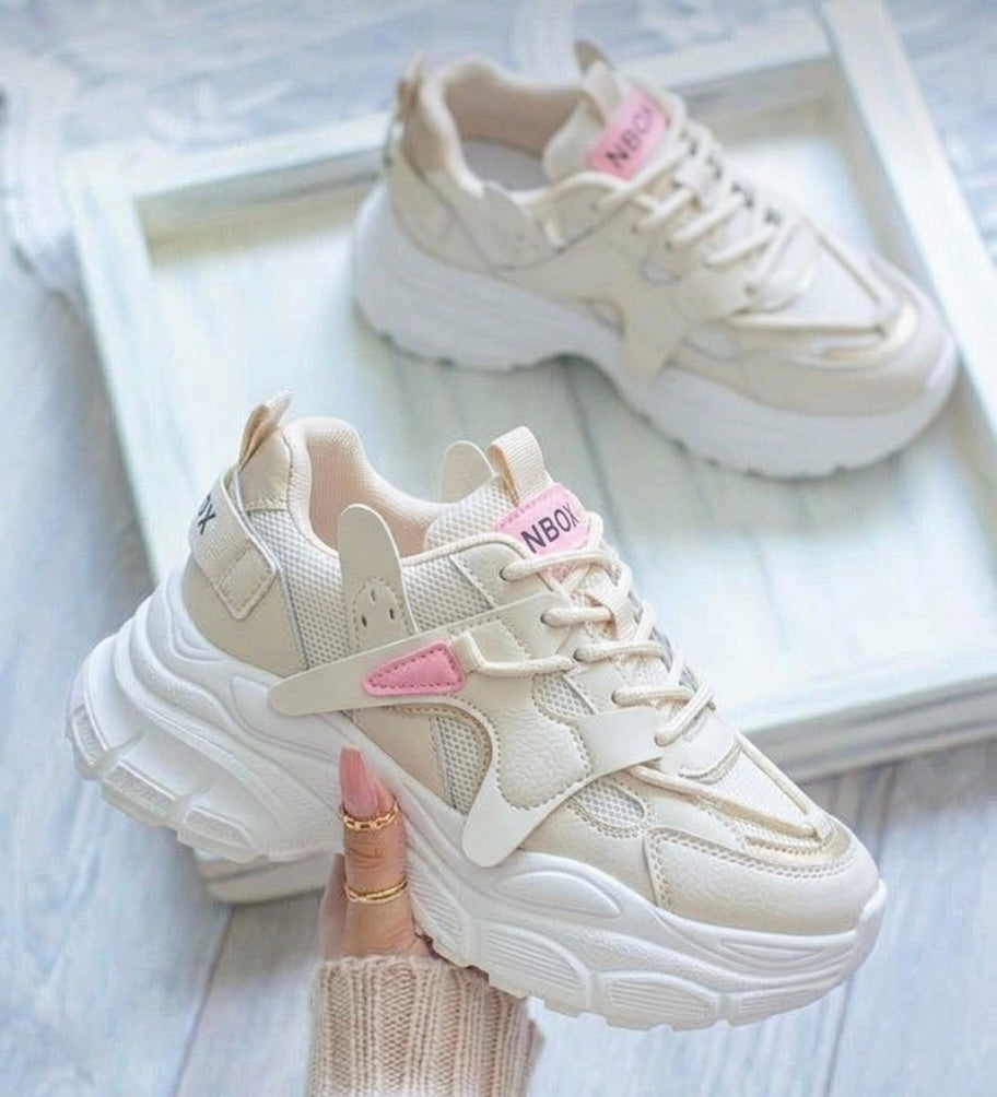 A pair of chunky platform sneakers in cream and white with pink accents, held by a hand with gold rings, with the other shoe resting on a white tray in the background.