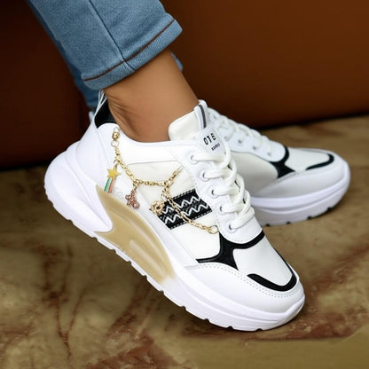 White sneakers with black accents and gold charms worn by a person on a brown surface.